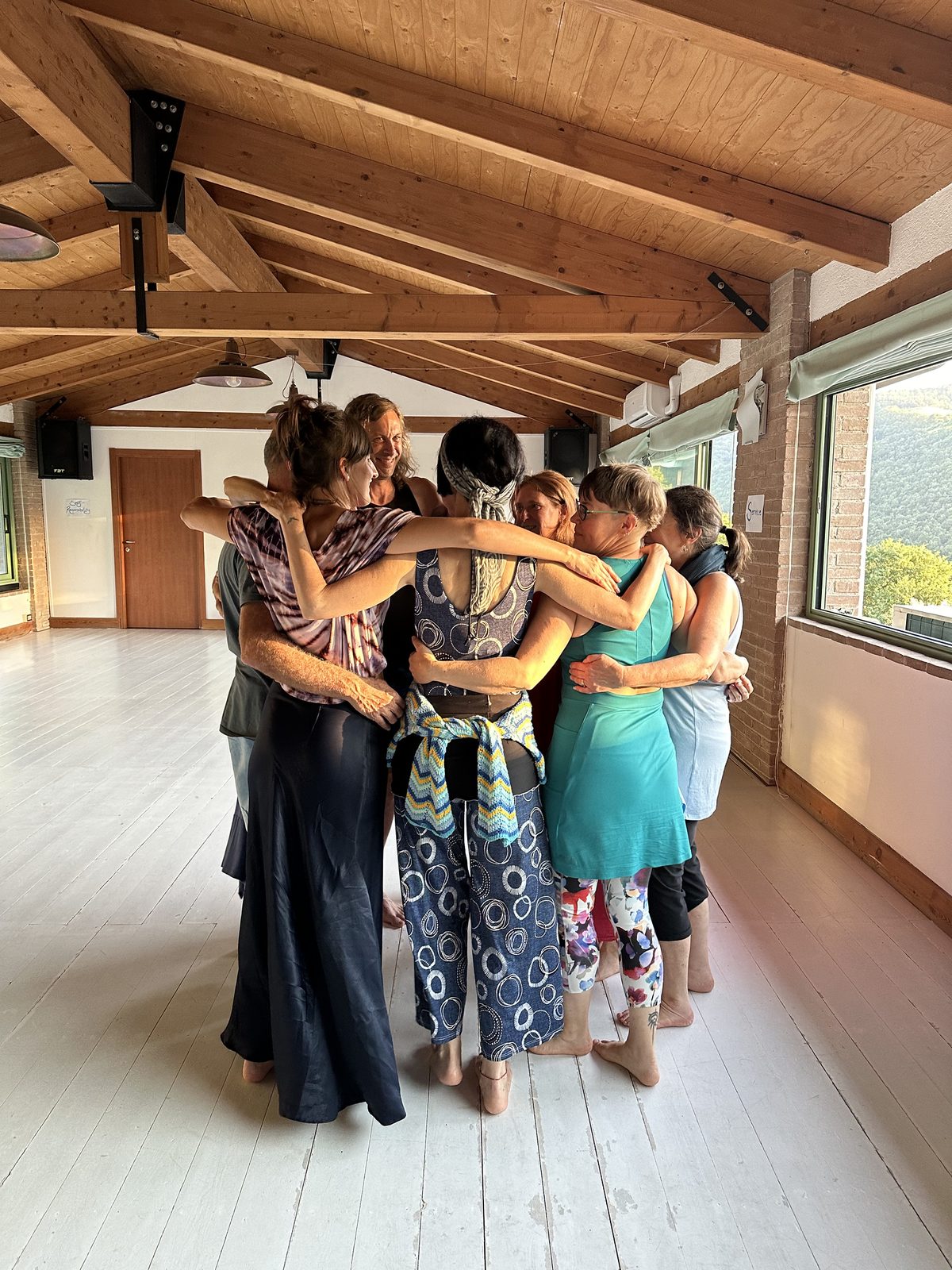 A circle of dancers standing close in a huddle, arms around one another, in a timber-beamed studio