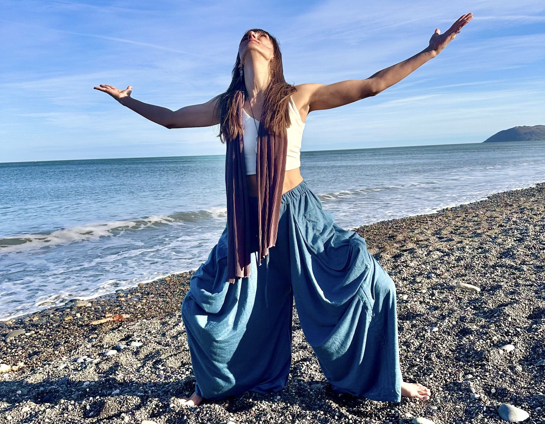 Gita barefoot on a stony beach, arms spread wide and face lifted to the sun, the sea behind her